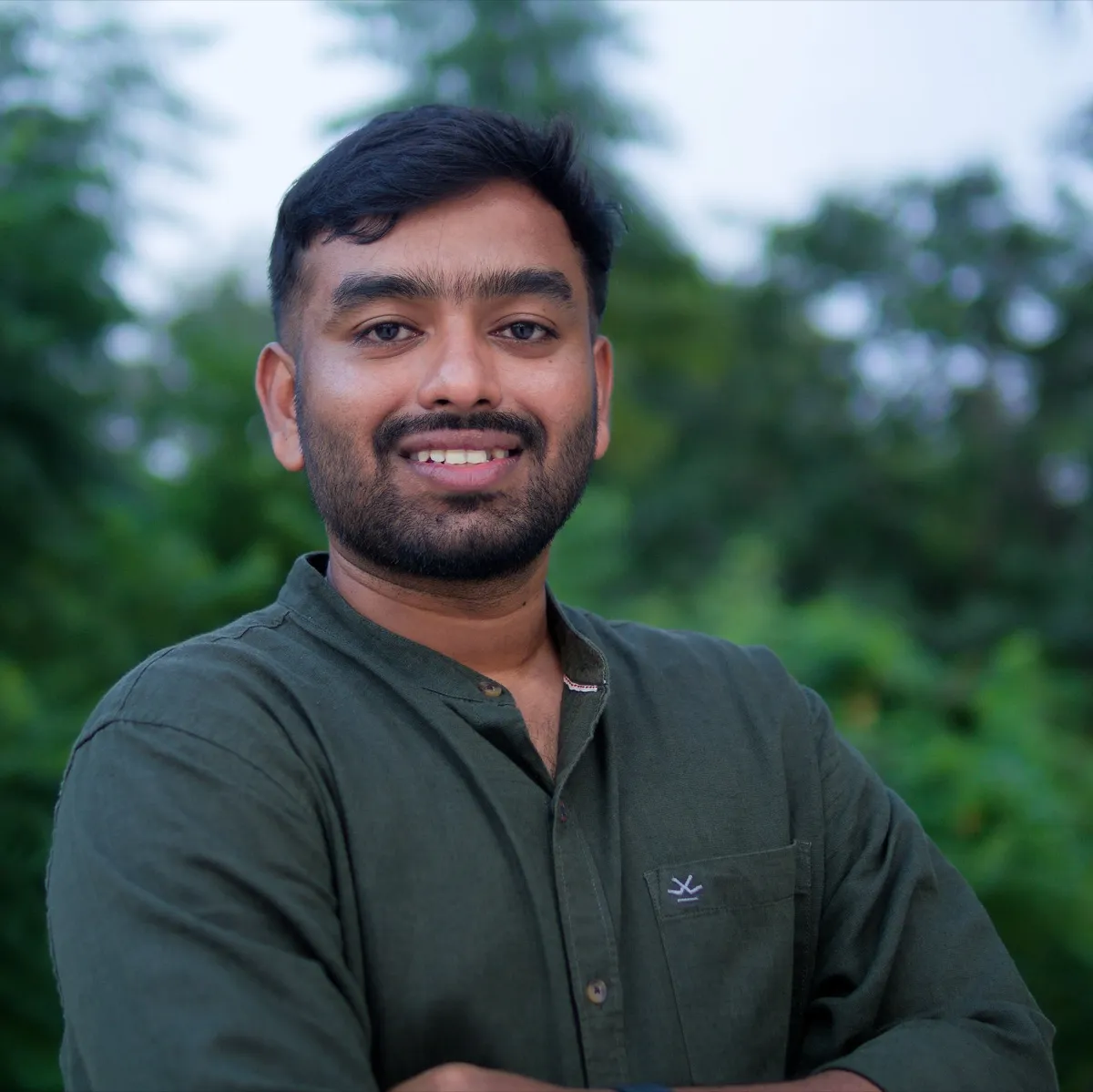 Isaac Solomon smiling, wearing a dark green shirt, greenery behind him